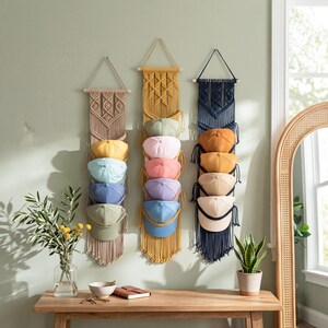 May include: Three macrame hat organizers in beige, yellow, and navy blue, each holding a stack of baseball caps in various colors. The organizers hang on a light green wall above a wooden table with a vase and a potted plant.