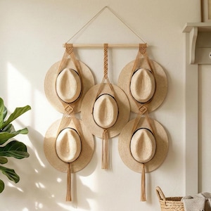 Puede incluir: Un perchero de macramé con seis sombreros de paja. Los sombreros están suspendidos de una barra de madera con cuerdas de color natural. El perchero está montado en una pared blanca. Cada sombrero tiene una banda negra.