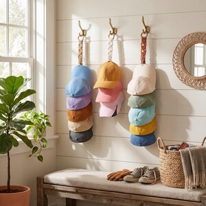 May include: A selection of baseball caps in shades of blue, pink, and beige, suspended from decorative hooks on a white wall. The caps are displayed in stacks. A bench, mirror, and basket are also visible in the room.
