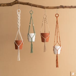 May include: Four macrame plant hangers with different coloured pots. The hangers are made of natural cotton rope and have a simple, minimalist design. The pots are in shades of brown, white, and green.