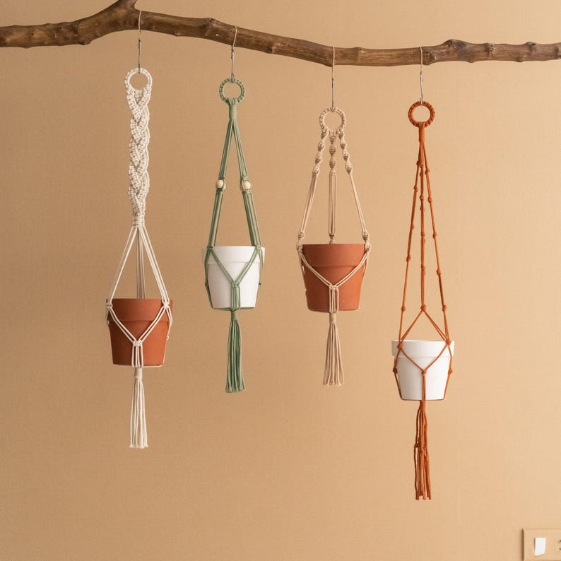 May include: Four macrame plant hangers with different coloured pots. The hangers are made of natural cotton rope and have a simple, minimalist design. The pots are in shades of brown, white, and green.