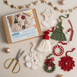 May include: A DIY kit for making macrame ornaments. The kit includes a variety of colours and patterns, including a snowflake, a wreath, a gnome, a candy cane, and a Christmas tree. The kit also includes a pair of scissors and a box with the text "DIY Kit Macrame Ornaments BeanDaikon".