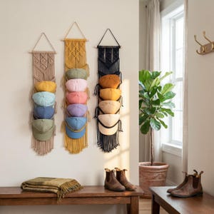 May include: Three macrame hat organizers in beige, yellow, and black, each holding six baseball caps in various colors. The organizers hang on a light-colored wall, with a wooden bench and boots in the foreground.