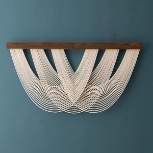 May include: A white macrame wall hanging with a wooden frame. The macrame is made of multiple strands of cord that are woven together to create a geometric pattern.