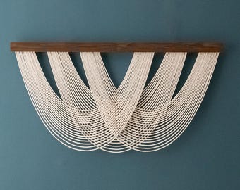 May include: A wall hanging featuring a horizontal dark brown wooden bar from which multiple off-white cotton cords are suspended. The cords are arranged in a series of overlapping, symmetrical U-shaped loops that create a layered, curved pattern.