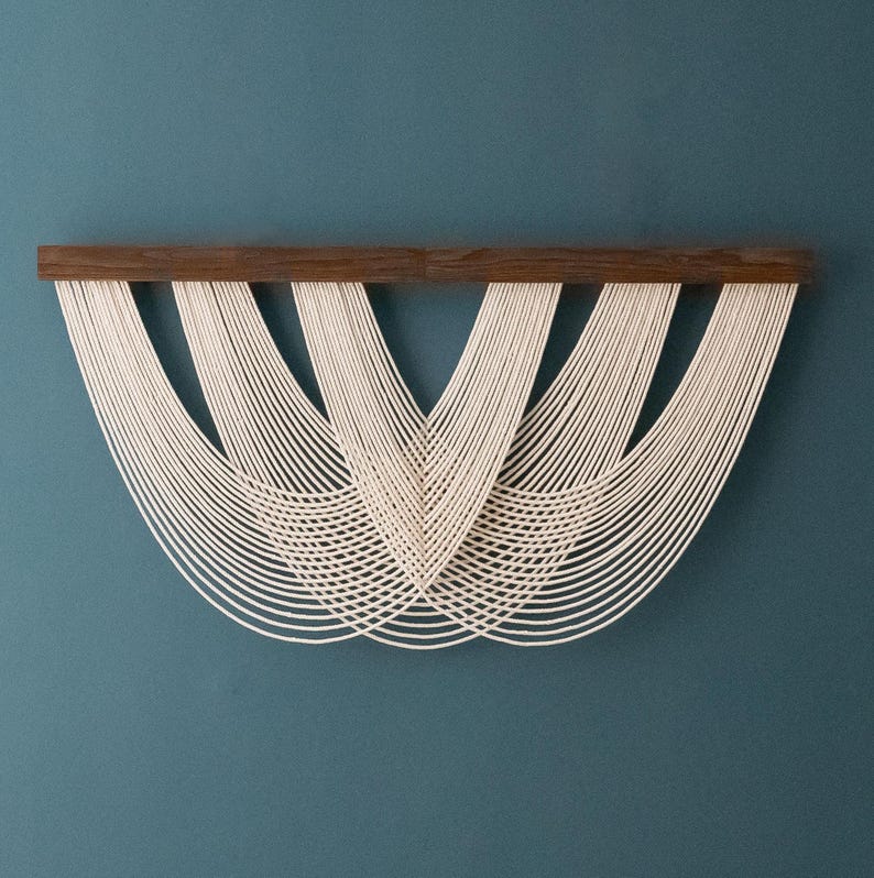 May include: A wall hanging featuring a horizontal dark brown wooden bar from which multiple off-white cotton cords are suspended. The cords are arranged in a series of overlapping, symmetrical U-shaped loops that create a layered, curved pattern.