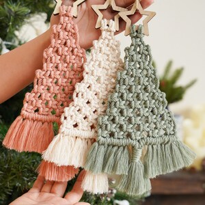 May include: Three macrame Christmas tree ornaments in pink, white, and green with wooden star toppers.