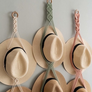 May include: Three macrame hat hangers in cream, sage green, and dusty rose, each featuring a wooden ring at the top and intricate knotted cord designs. These hangers hold straw hats with black bands, displaying a vertical arrangement of headwear.