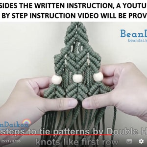 Puede incluir: Primer plano de un tapiz de macram&eacute; verde con cuentas de madera blancas. El macram&eacute; tiene forma de tri&aacute;ngulo y tiene flecos en la parte inferior. El texto "BeanDaikon" y "beandaikon.etsy" es visible en la imagen.