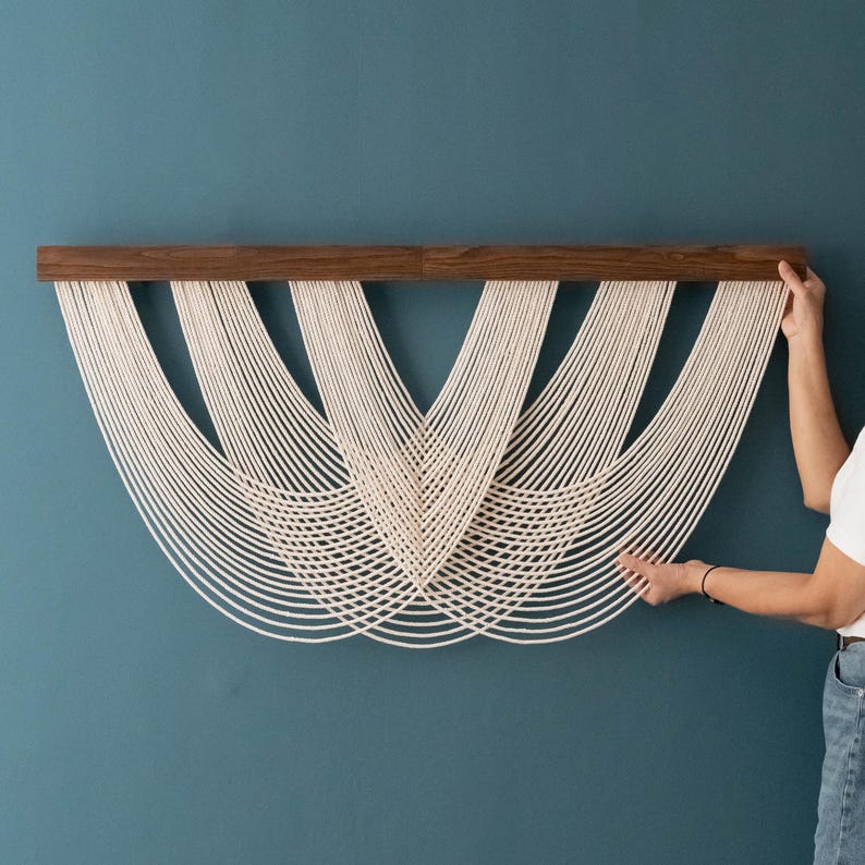 May include: A wall hanging featuring a horizontal dark brown wooden bar from which multiple off-white cotton cords are suspended. The cords are arranged in a series of overlapping, symmetrical U-shaped loops that create a layered, curved pattern.