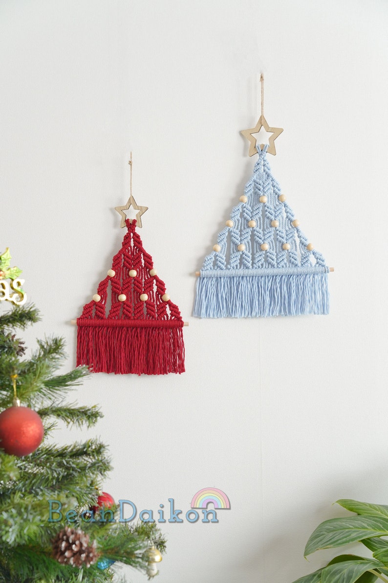 Puede incluir: Dos decoraciones de pared de macram&eacute; en forma de &aacute;rbol de Navidad, una roja y otra azul claro, con cuentas de madera y una estrella de madera en la parte superior.