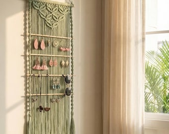 May include: A sage green macrame wall hanging organizer featuring four horizontal wooden dowels for displaying jewelry. The piece includes intricate knotted patterns at the top, long fringe at the bottom, and holds various earrings, necklaces, and sunglasses.