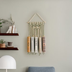 May include: A green and white macrame wall hanging with a wooden dowel at the top. Three books are hanging from the bottom of the macrame, with the titles "LOEWE", "VOGUE", and "talton" visible.