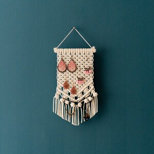 May include: A white macrame wall hanging with a fringe border. The hanging has multiple loops for holding earrings and necklaces. The hanging features pink and brown earrings and a brown necklace.