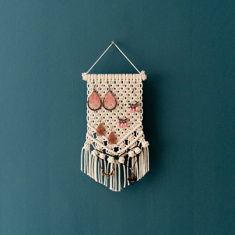 May include: A white macrame wall hanging with a fringe border. The hanging has multiple loops for holding earrings and necklaces. The hanging features pink and brown earrings and a brown necklace.