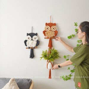 Peut inclure: Deux tentures murales en macram&eacute; en forme de hibou, l'une orange et l'autre grise, avec des accents de fil blanc. Un pot suspendu avec une plante verte se trouve au centre. Le pot est en macram&eacute; avec un pot en c&eacute;ramique blanc.