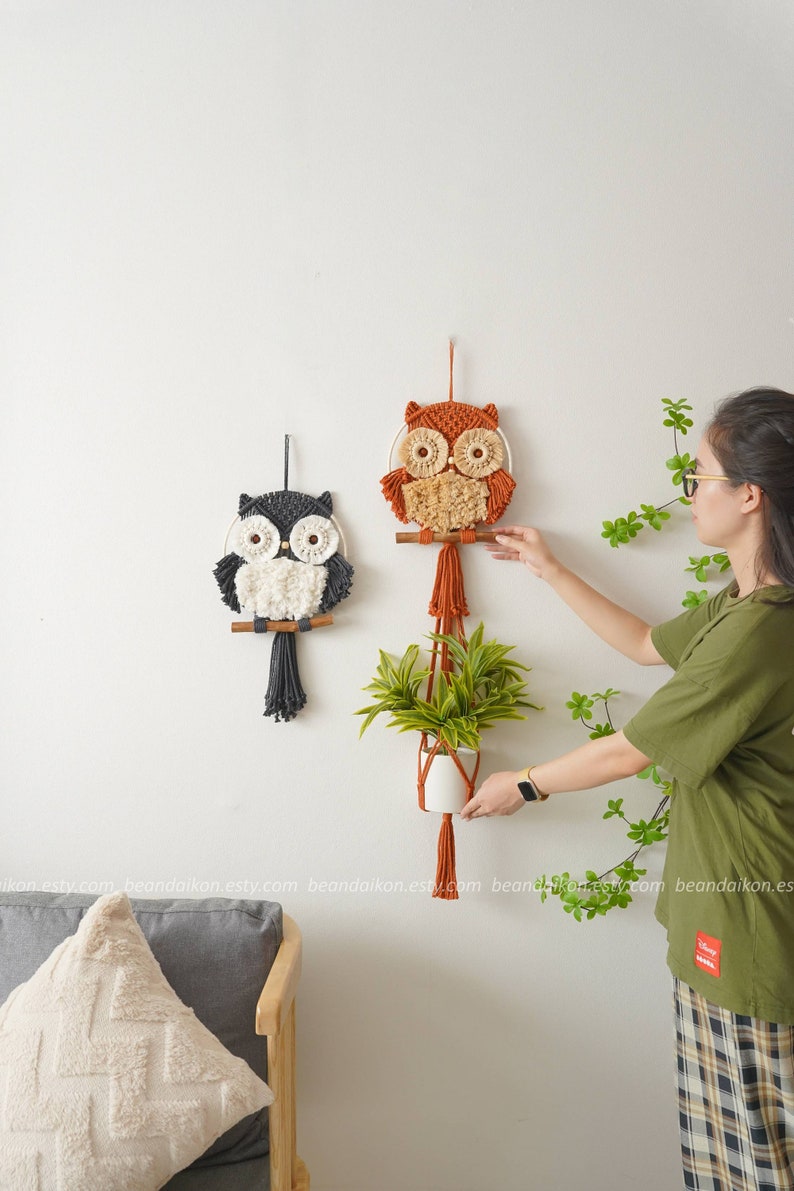 Peut inclure: Deux tentures murales en macram&eacute; en forme de hibou, l'une orange et l'autre grise, avec des accents de fil blanc. Un pot suspendu avec une plante verte se trouve au centre. Le pot est en macram&eacute; avec un pot en c&eacute;ramique blanc.