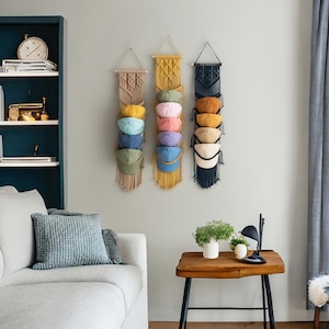May include: Three macrame hat organizers in beige, yellow, and navy blue, each holding several baseball caps in various colors. The organizers are hanging on a light gray wall, with a sofa and side table in the foreground.