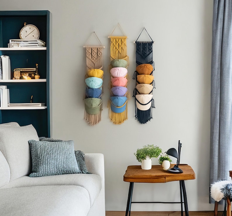 May include: Three macrame hat organizers in beige, yellow, and navy blue, each holding several baseball caps in various colors. The organizers are hanging on a light gray wall, with a sofa and side table in the foreground.