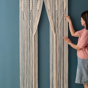 K&ouml;nnte beinhalten: Ein wei&szlig;er Makramee-Vorhang mit geometrischem Muster h&auml;ngt an einer blaugr&uuml;nen Wand. Der Vorhang besteht aus dickem Baumwollschnur und hat oben eine Holzstange.