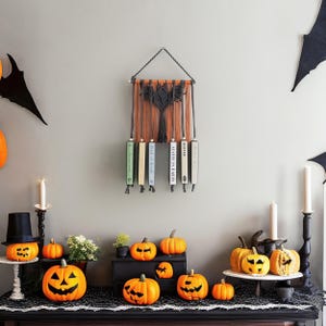 May include: A black macrame wall hanging with a geometric design and five books hanging from it. The books are titled "Butter", "Mail is in Turin", "Book of Blood", "The Book of Lies", and "The Book of Lies". There are several orange pumpkins on a black table in front of the wall hanging.