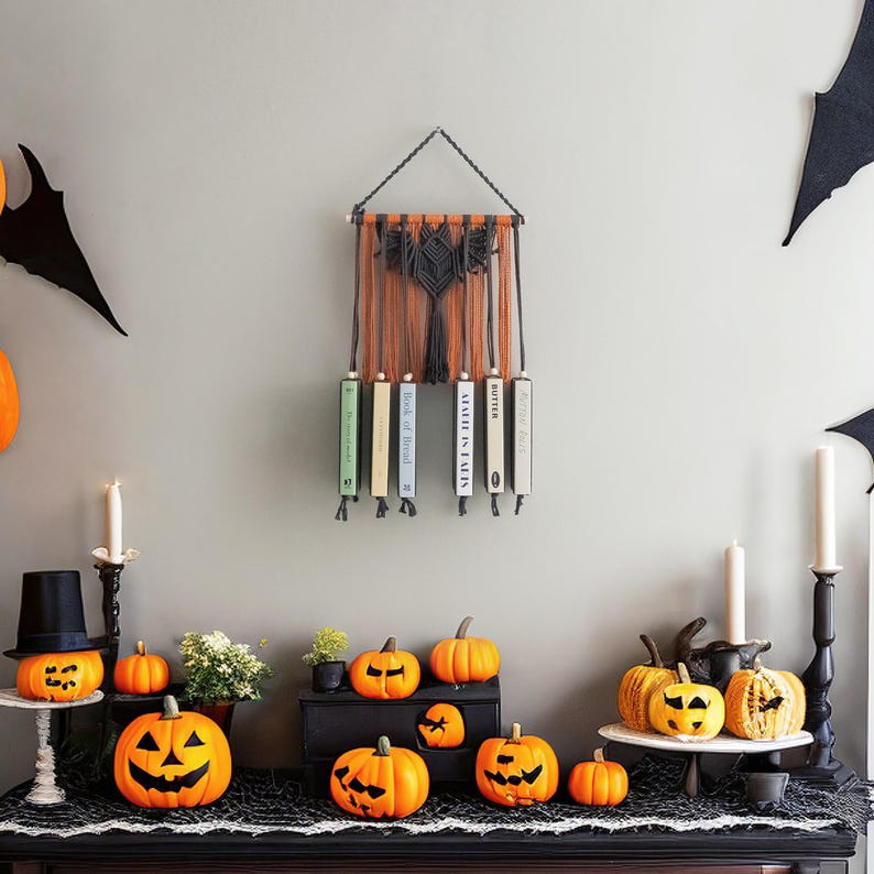 May include: A black macrame wall hanging with a geometric design and five books hanging from it. The books are titled "Butter", "Mail is in Turin", "Book of Blood", "The Book of Lies", and "The Book of Lies". There are several orange pumpkins on a black table in front of the wall hanging.