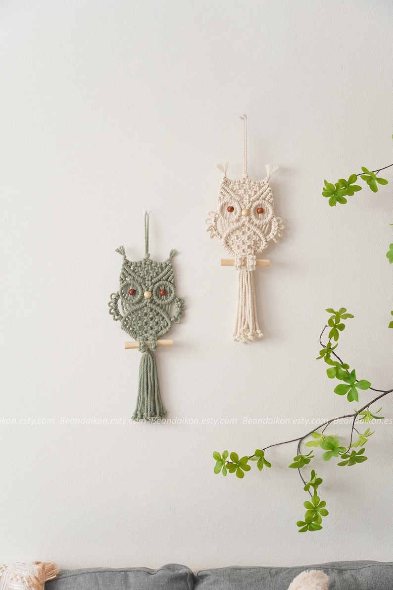 Peut inclure: Deux d&eacute;corations murales en macram&eacute; en forme de hibou. L'une est vert sauge, l'autre blanc cass&eacute;. Les deux ont des chevilles en bois et des yeux perl&eacute;s. Les hiboux sont accroch&eacute;s &agrave; un mur blanc, avec du feuillage vert en arri&egrave;re-plan.