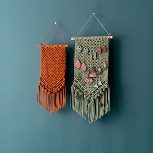 May include: Two macrame wall hangings, one in a green color and one in a burnt orange color. The green wall hanging has a variety of colorful earrings hanging from it. Both wall hangings have fringe at the bottom.