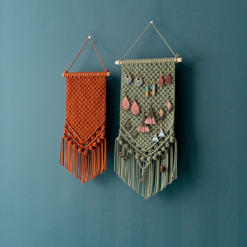 May include: Two macrame wall hangings, one in a green color and one in a burnt orange color. The green wall hanging has a variety of colorful earrings hanging from it. Both wall hangings have fringe at the bottom.