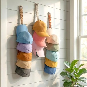 May include: A selection of baseball caps in shades of blue, pink, and yellow are arranged on a wall. The caps are suspended from decorative rope hangers, providing a fashionable and tidy storage option. The wall is painted white with horizontal panelling.