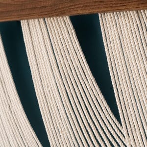 May include: A wall hanging featuring numerous off-white cotton cords suspended from a horizontal wooden dowel. The cords are arranged in vertical sections, creating a series of parallel lines that hang downward from the dark brown wood support.