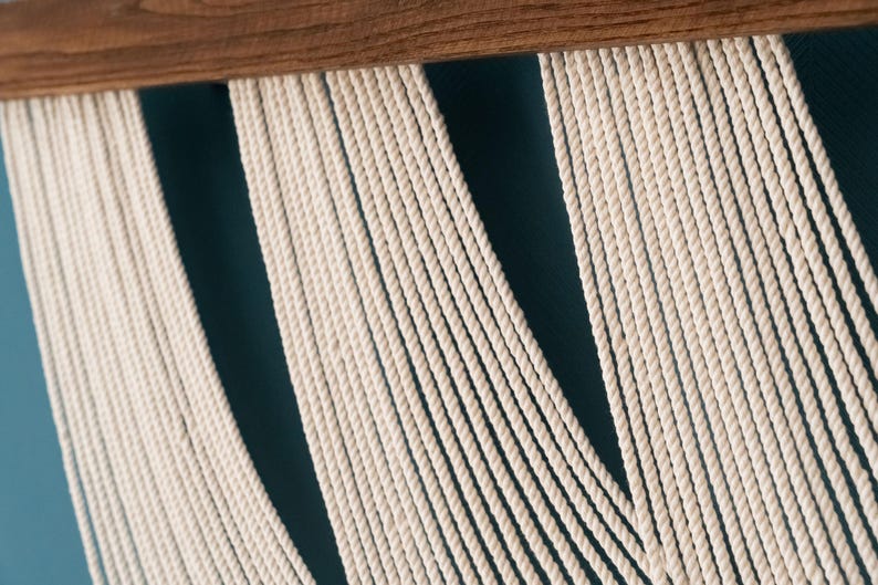 May include: A wall hanging featuring numerous off-white cotton cords suspended from a horizontal wooden dowel. The cords are arranged in vertical sections, creating a series of parallel lines that hang downward from the dark brown wood support.