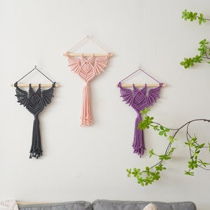 May include: Three macrame wall hangings in the shape of bats with wooden dowels at the top. The bats are in pink, purple, and grey.