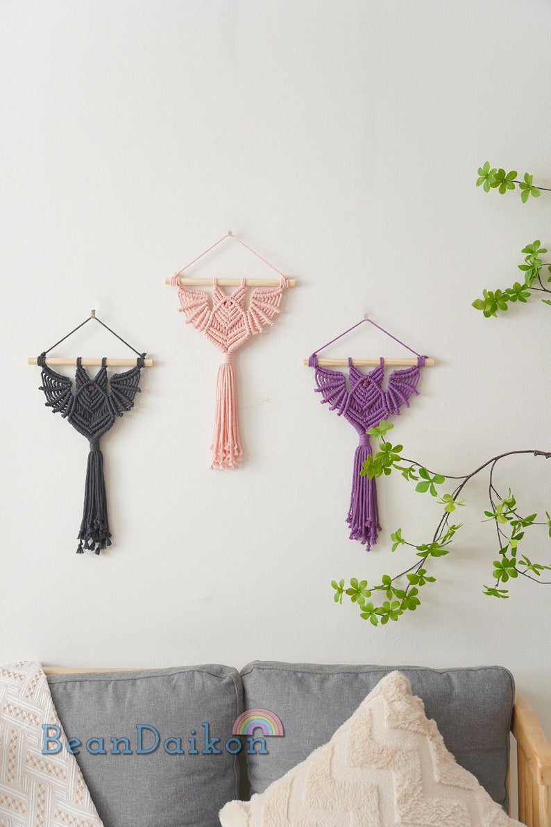 May include: Three macrame wall hangings in the shape of bats with wooden dowels at the top. The bats are in pink, purple, and grey.