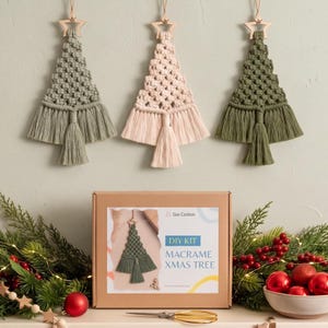 Puede incluir: Tres adornos de pared de macramé con forma de árbol de Navidad en verde salvia, rosa pálido y verde oliva, rematados con una estrella de madera. Debajo, una caja de cartón marrón con la etiqueta Sisi Cotton DIY KIT MACRAME XMAS TREE.