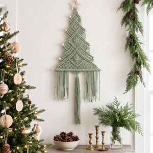May include: A sage green macrame wall hanging shaped like a Christmas tree, featuring intricate knotted patterns, light wooden beads along the edges, a wooden star at the top, and a long fringe at the bottom suspended from a wooden dowel.