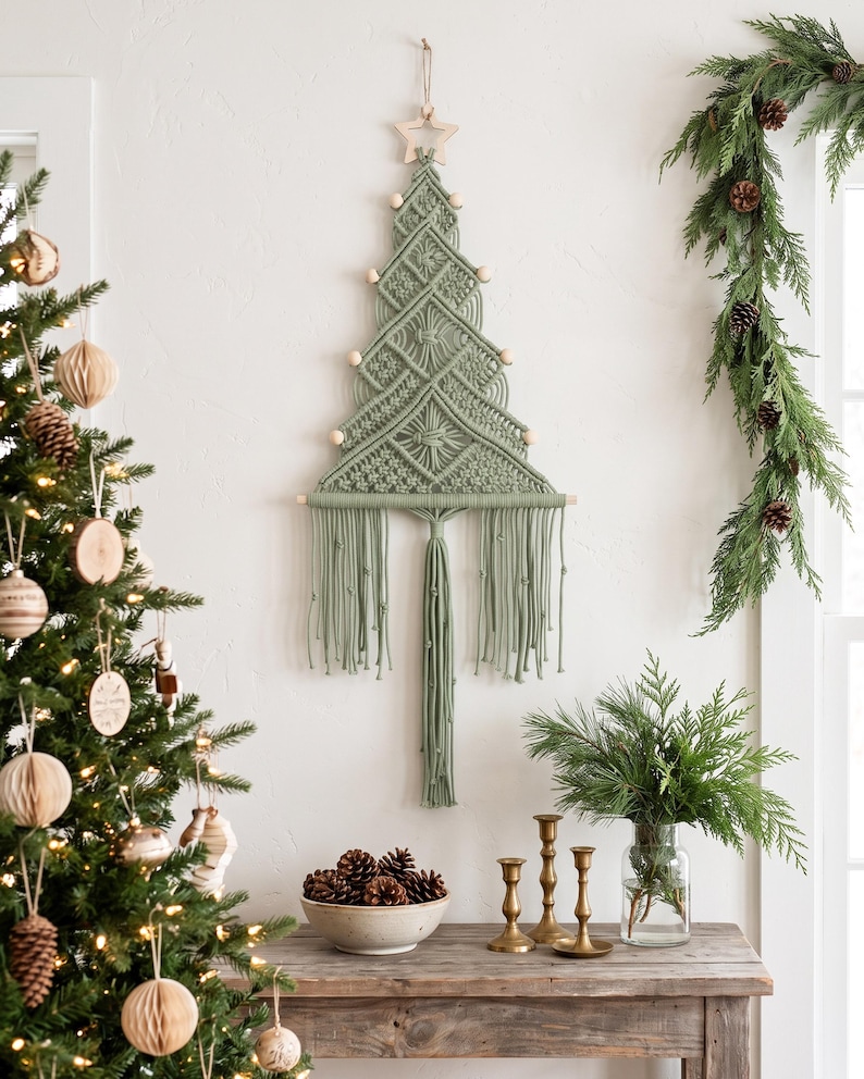 May include: A sage green macrame wall hanging shaped like a Christmas tree, featuring intricate knotted patterns, light wooden beads along the edges, a wooden star at the top, and a long fringe at the bottom suspended from a wooden dowel.