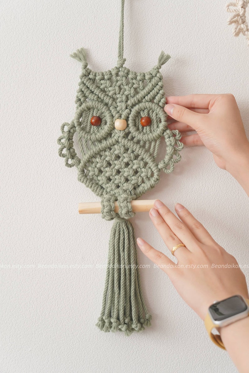 Peut inclure: D&eacute;coration murale en macram&eacute; en forme de hibou vert sauge. Le hibou a des yeux en perles de bois et un perchoir en cheville de bois. Le hibou est suspendu par une boucle en haut et a une gland en bas. Fait &agrave; la main.
