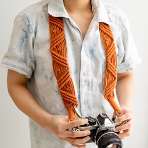 May include: A person wearing a light blue shirt with a tie-dye pattern is holding a silver Nikon camera with a brown macrame camera strap. The camera has a black lens and a black leather grip.