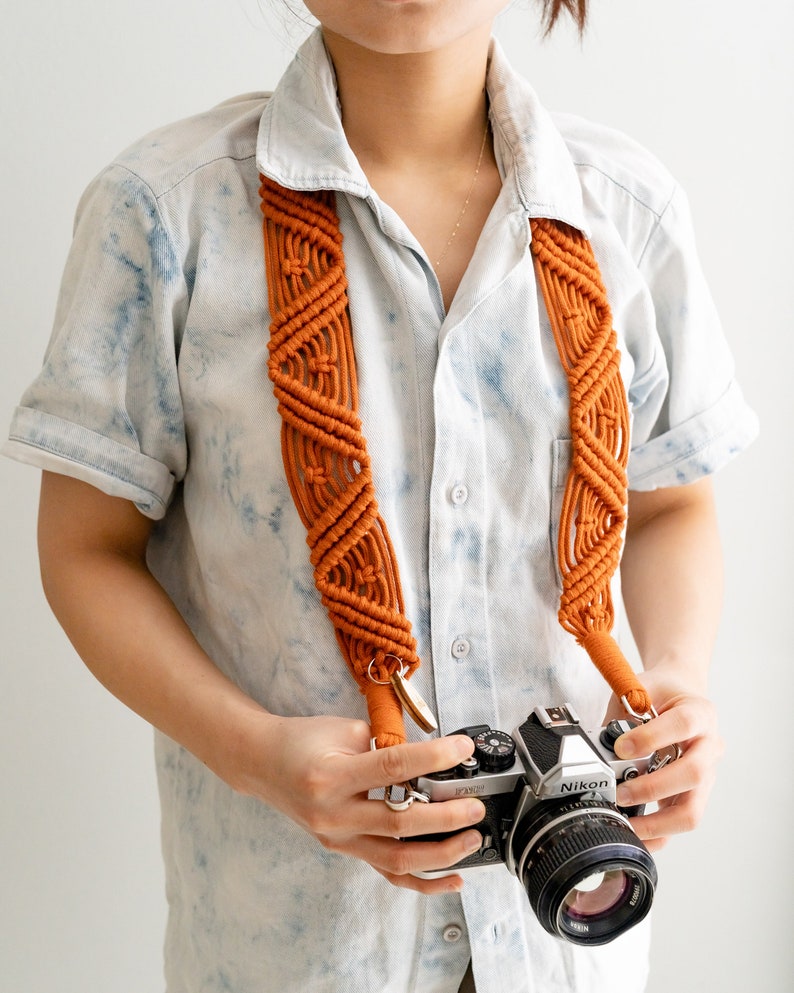 May include: A person wearing a light blue shirt with a tie-dye pattern is holding a silver Nikon camera with a brown macrame camera strap. The camera has a black lens and a black leather grip.