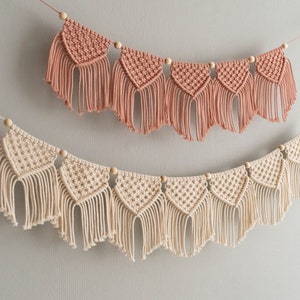 Puede incluir: Dos banderines de macramé, uno en color rosa empolvado y otro en crema, están suspendidos sobre un fondo gris. Cada banderín presenta una serie de formas triangulares con flecos y cuentas de madera, creando una estética bohemia decorativa.