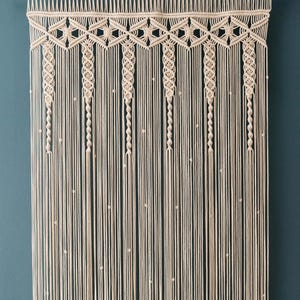 以下が含まれることがあります： 幾何学模様とビーズの糸でできたマクラメの壁掛け。ナチュラルカラーのコードで作られており、高さは約90cmです。