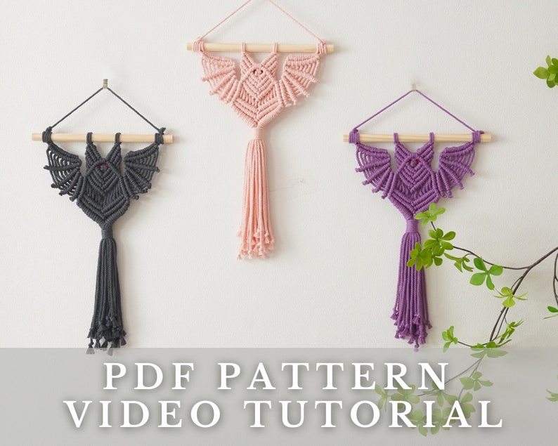 May include: Three macrame wall hangings in the shape of bats, one in pink, one in purple, and one in grey. The hangings are made with a knotting technique and have tassels at the bottom. The text "PDF PATTERN VIDEO TUTORIAL" is at the bottom of the image.
