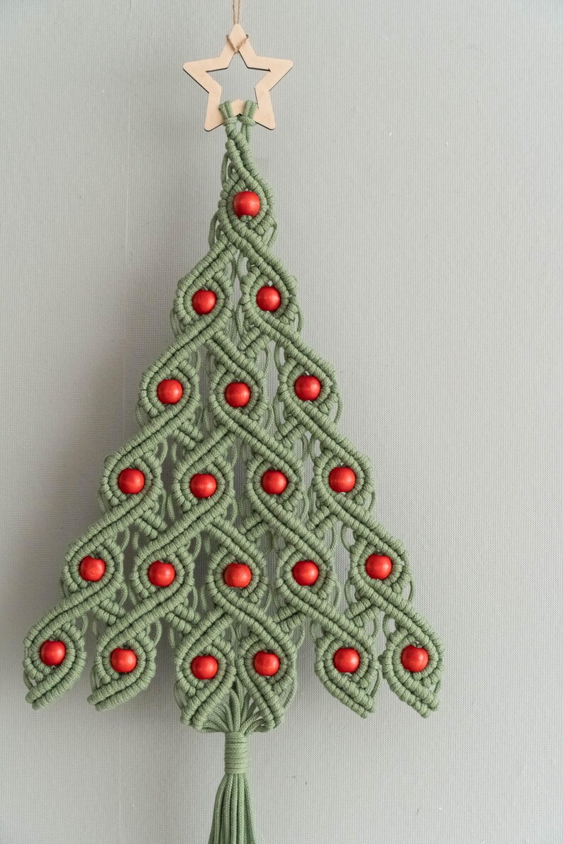 K&ouml;nnte beinhalten: Ein gr&uuml;ner Makramee-Weihnachtsbaum-Wandbehang mit roten Holzperlen und einem Holzstern an der Spitze.