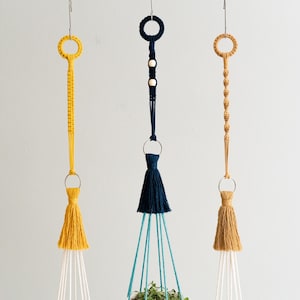 May include: Three macrame plant hangers with wooden beads and tassels. The hangers are in blue, yellow, and brown.