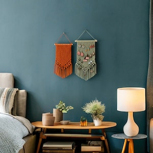 May include: Two macrame wall hangings, one orange and one green, with fringe, hanging on a teal wall. The green wall hanging has a small pocket for holding jewelry.