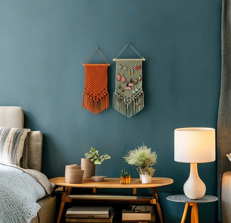 May include: Two macrame wall hangings, one orange and one green, with fringe, hanging on a teal wall. The green wall hanging has a small pocket for holding jewelry.