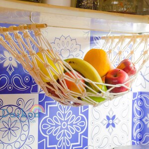 May include: A white rope hammock hanging from a shelf, filled with fruit, including a yellow banana, a green apple, a red apple, and an orange. The hammock is hanging in front of a blue and white tiled wall.