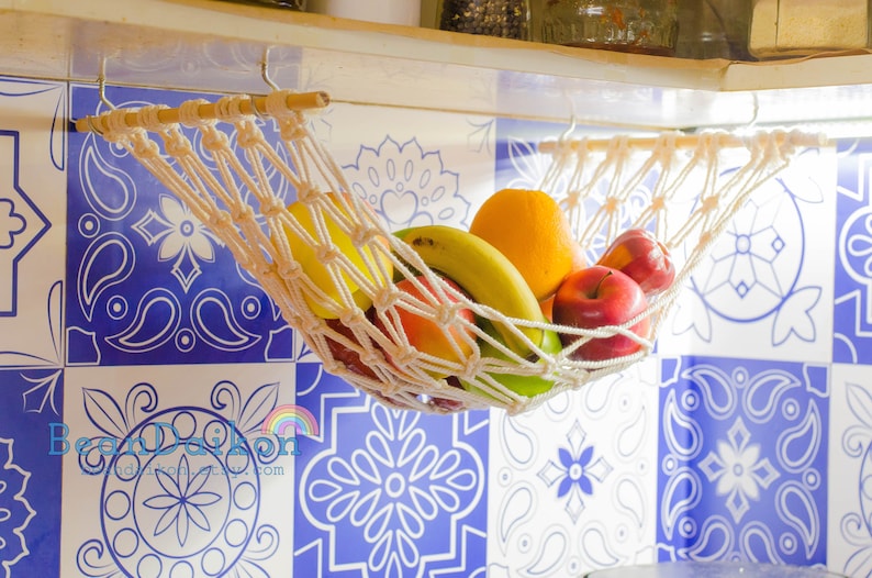 May include: A white rope hammock hanging from a shelf, filled with fruit, including a yellow banana, a green apple, a red apple, and an orange. The hammock is hanging in front of a blue and white tiled wall.