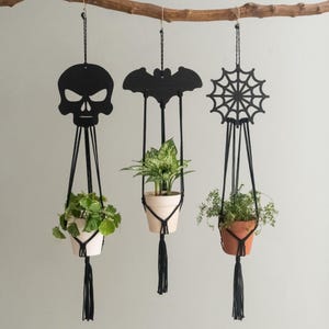 May include: Three Halloween-themed macrame plant hangers suspended from a branch. The black silhouettes include a skull, a bat, and a spiderweb. Each hanger supports a small potted plant with green foliage, creating a spooky yet stylish decor.